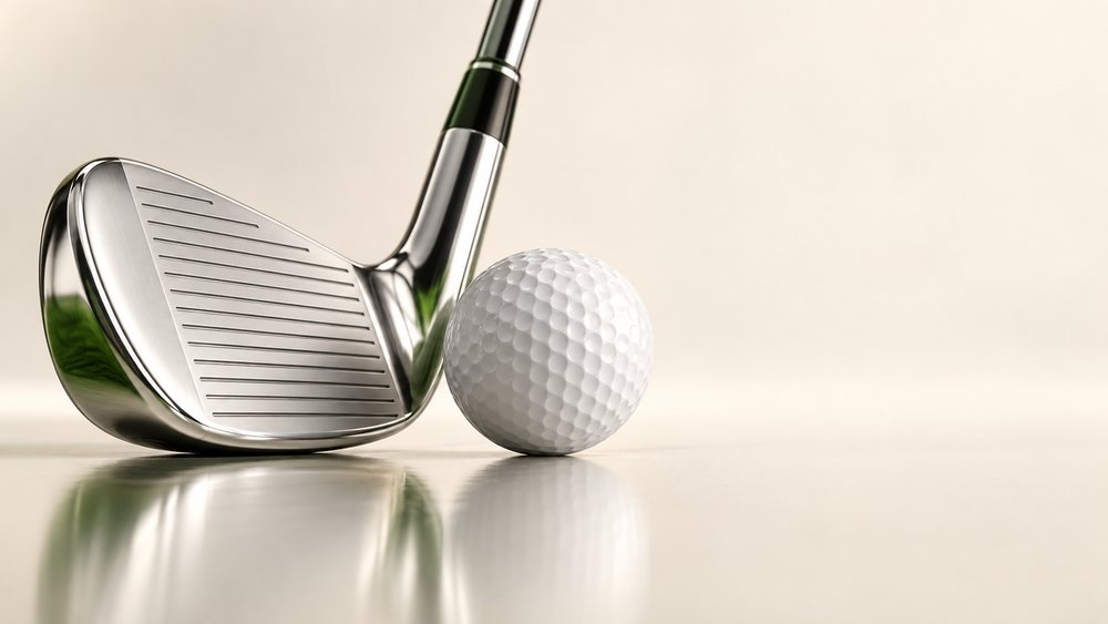 A chrome iron resting behind a white golf ball on a pale studio surface